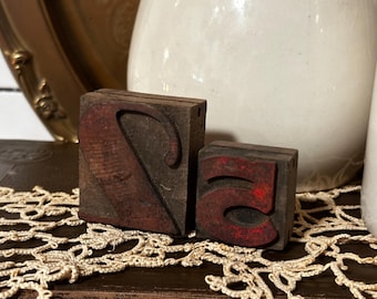 Op de afbeelding: Twee vintage houten drukblokken, één met de letter "S" en de andere met het cijfer "5". De blokken zijn donkerbruin met rode inktresten, wat duidt op leeftijd en gebruik. Ze liggen op een decoratief kanten kleedje.