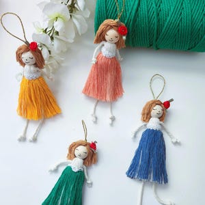 May include: Handmade doll ornaments with yarn hair, red flower accents, and colorful skirts in yellow, peach, green, and blue. Each doll has a white top and dangling legs. A spool of green yarn and white flowers are in the background.