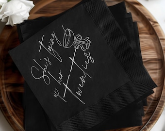 May include: Black paper napkins with white text that reads "She's tying up her twenties". A white illustration of a cocktail glass with a straw is above the text.