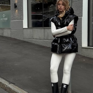 Könnte beinhalten: Eine schwarze, glänzende Steppweste mit Kapuze, getragen über einem weißen Langarmshirt und einer weißen Leggings. Das Outfit wird durch schwarze Gummistiefel mit einem weißen Doppel-C-Logo am Schaft ergänzt, was einen starken Kontrast erzeugt.