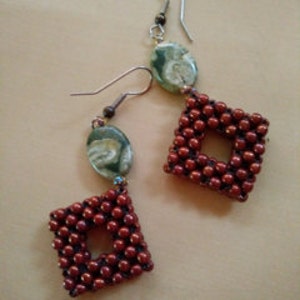 May include: A pair of handcrafted earrings featuring square, open-center designs. The earrings are made with small, reddish-brown beads and are accented with oval-shaped green and white stone beads. The ear wires are silver-colored.