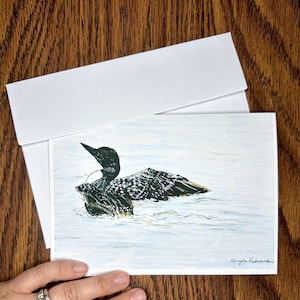 Puede incluir: Una ilustración en blanco y negro de un colimbo con ojos rojos nadando en el agua. El colimbo tiene las alas extendidas y mira hacia la derecha. La firma del artista, "Angela Kishwaukee", está en la esquina inferior derecha de la tarjeta.