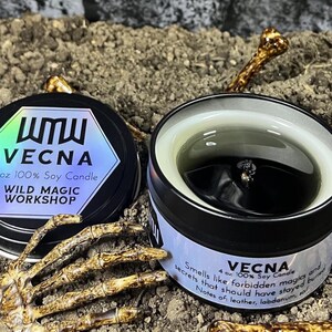 May include: Two black 4 oz soy candles from Wild Magic Workshop. One candle is open, revealing a black wax interior. The closed candle has a label with the text "VECNA" and "4 oz 100% Soy Candle". The candles are surrounded by a skeleton hand and bone fragments.