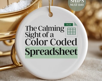 May include: A white, round Christmas ornament with the text "The Calming Sight of a Color Coded Spreadsheet." The ornament has a green spreadsheet graphic and is hanging from a gold string. A small gift box and a miniature Christmas tree are in the background.