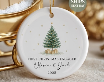 May include: A white ceramic Christmas ornament with a green Christmas tree design, gold stars, and gift boxes. The text reads "FIRST CHRISTMAS ENGAGED Olivia & Jack 2025". A gold string is attached for hanging.