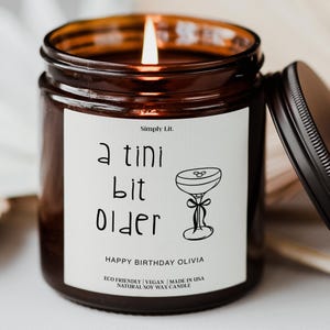 May include: A lit candle in a brown glass jar with a black lid. The label reads "a tini bit older" with a cocktail glass illustration. The text "HAPPY BIRTHDAY OLIVIA" is also on the label. The candle is made with natural soy wax.