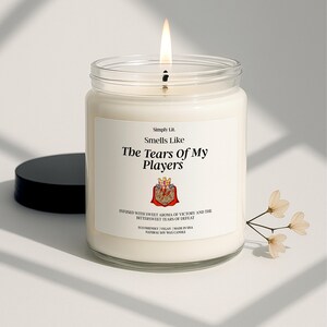 May include: A lit candle in a clear glass jar with a black lid. The label reads "Smells Like The Tears Of My Players." The candle is white with a burning wick. A sprig of white flowers is next to the candle.