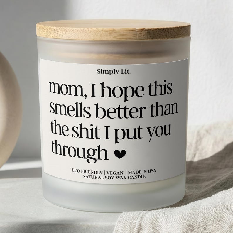 May include: A frosted glass candle with a wooden lid. The label reads "mom, I hope this smells better than the shit I put you through" in black text. The candle is labeled "Eco Friendly | Vegan | Made in USA | Natural Soy Wax Candle."
