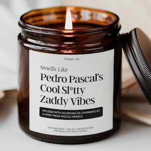May include: A lit soy wax candle in a brown glass jar with a black lid. The label reads "Smells Like Pedro Pascal's Cool Sl*tty Zaddy Vibes." The candle is eco-friendly, vegan, and made in the USA.