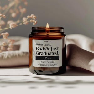 May include: A lit candle in a brown glass jar. The label reads "Smells Like A Baddie Just Graduated. Olivia. Class of 2026." The candle is eco-friendly, vegan, and made with natural soy wax.