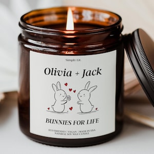 May include: A lit soy wax candle in a brown glass jar with a black lid. The label reads "Olivia + Jack" above a graphic of two bunnies and the text "Bunnies For Life". The candle is eco-friendly, vegan, and made in the USA.