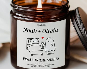 May include: A lit soy wax candle in a brown glass jar with a black lid. The label reads "Noah + Olivia" above a cartoon of a ghost and a person in bed, with the text "FREAK IN THE SHEETS".