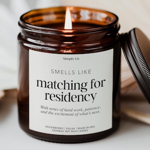 May include: A lit soy wax candle in a brown glass jar with a black lid. The white label reads Simply Lit. SMELLS LIKE matching for residency. With notes of hard work, patience, and the excitement of what's next. ECO FRIENDLY | VEGAN | MADE IN USA.