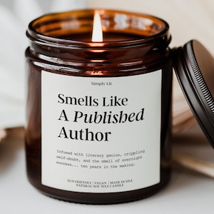 May include: A lit, brown glass jar candle with a black lid. The candle has a white label with the text "Smells Like A Published Author". The candle is made of natural soy wax and is eco-friendly and vegan.