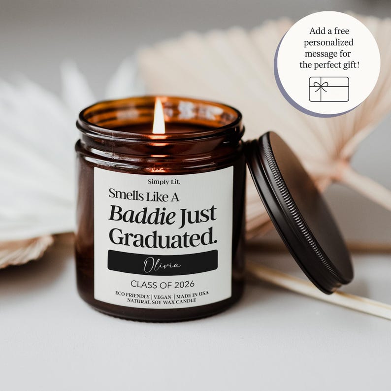 May include: A lit, brown glass jar candle with a black lid, featuring the text "Smells Like A Baddie Just Graduated." The candle includes the name "Olivia" and "CLASS OF 2026." The candle is labeled as eco-friendly, vegan, and made in the USA.