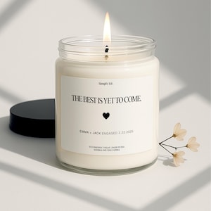 May include: A lit, white candle in a clear glass jar with a black lid. The label reads "THE BEST IS YET TO COME" with a heart and the names "EMMA + JACK ENGAGED 2.22.2025". A sprig of flowers is next to the candle.
