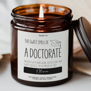 May include: A lit brown glass jar candle with a black lid. The candle has a white label with the text "The Sweet Smell of a Doctorate" and "Olivia". The candle is made of natural soy wax.