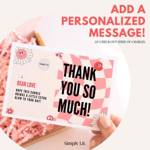 May include: A greeting card with the text "THANK YOU SO MUCH!" in black on a pink and white checkered background. The card also includes the text "DEAR LOVE" and "HOPE THIS CANDLE BRINGS A LITTLE EXTRA GLOW TO YOUR DAY!" with flower and smiley face graphics. The card is in a gift box.
