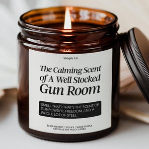 May include: A lit brown glass jar candle with a black lid. The label reads "The Calming Scent of A Well Stocked Gun Room." The text below says "Smell that? That's the scent of gunpowder, freedom, and a whole lot of steel." The candle is made of natural soy wax.