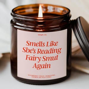 May include: A lit candle in a brown glass jar with a black lid. The white label features the text Simply Lit. and Smells Like She's Reading Fairy Smut Again in red serif font, with Eco Friendly, Vegan, Made in USA, Natural Soy Wax Candle below.
