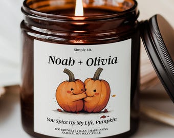 May include: A lit candle in a brown glass jar with a black lid. The label reads "Noah + Olivia" above an illustration of two hugging pumpkins. The text "You Spice Up My Life, Pumpkin" is below the pumpkins. The candle is made in the USA.