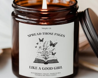 May include: A lit candle in a brown glass jar with a black lid. The white label features the text Simply Lit. SPREAD THOSE PAGES LIKE A GOOD GIRL, with a black and white illustration of an open book, wildflowers, and butterflies.