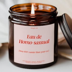 May include: A lit soy wax candle in a brown glass jar with a black lid. The label reads "Eau de Homo-sassual" in red text, with the tagline "Now 100% sassier than your ex." The candle is eco-friendly and vegan.
