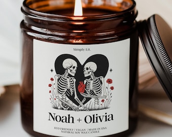 May include: A lit soy wax candle in a brown glass jar with a black lid. The label features a black heart with two skeletons, red flowers, and the names "Noah + Olivia." The text "Simply Lit" and candle details are also on the label.
