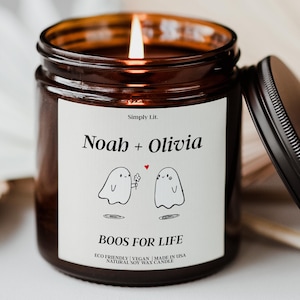 May include: A lit candle in a brown glass jar with a black lid. The white label features the text Simply Lit, Noah + Olivia, and BOOS FOR LIFE, with a graphic of two ghosts, one holding a flower, and a small red heart between them.