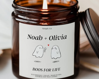 May include: A lit candle in a brown glass jar with a black lid. The white label features the text Simply Lit, Noah + Olivia, and BOOS FOR LIFE, with a graphic of two ghosts, one holding a flower, and a small red heart between them.