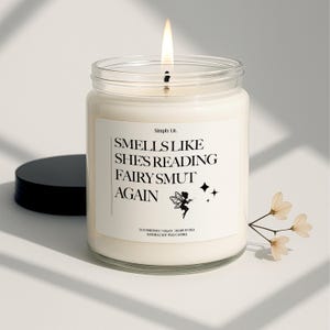 May include: A lit candle in a clear glass jar with a black lid. The label reads "Smells Like She's Reading Fairy Smut Again" with a fairy illustration. The candle is made with natural soy wax.