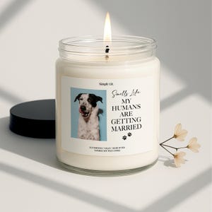 May include: A lit candle in a glass jar with a black lid. The label reads "Smells Like My Humans Are Getting Married" with a photo of a dog. The candle is a natural soy wax candle.