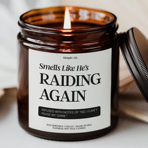 May include: A lit brown glass candle jar with a black lid, featuring a white label with the text "Smells Like He's RAIDING AGAIN." The candle is infused with notes of "No, I can't pause my game." It is an eco-friendly, vegan, natural soy wax candle.