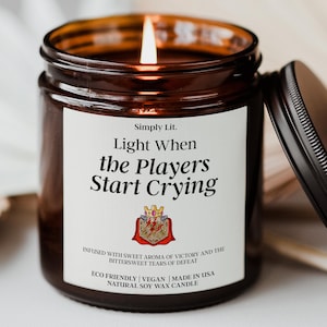 May include: A lit soy wax candle in a brown glass jar with a black lid. The white label features the text Simply Lit. Light When the Players Start Crying, an illustration of a crown atop a twenty-sided die with a red cape, and descriptive text.