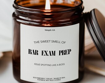 May include: A lit candle in a brown glass jar with a black lid. The white label reads Simply Lit. THE SWEET SMELL OF BAR EXAM PREP, ISSUE SPOTTING LIKE A BOSS, ECO FRIENDLY | VEGAN | MADE IN USA NATURAL SOY WAX CANDLE in black text.