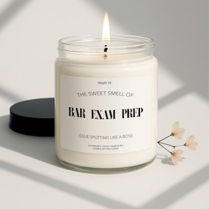 May include: A lit candle in a clear glass jar with a black lid. The label reads "THE SWEET SMELL OF BAR EXAM PREP" and "ISSUE SPOTTING LIKE A BOSS". A small sprig of flowers is next to the candle. The candle is made in the USA.