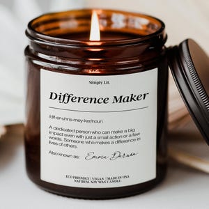 May include: A lit soy wax candle in a brown glass jar with a black lid. The label reads "Difference Maker" with a definition and the name "Emma Daruin." The candle is eco-friendly, vegan, and made in the USA.
