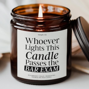 May include: A lit brown glass jar candle with a black lid. The candle has a white label with the text "Whoever Lights This Candle Passes the BAR EXAM". The candle is labeled as eco-friendly, vegan, and made in the USA.