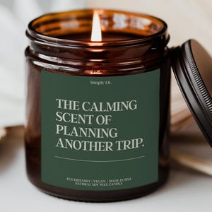 May include: A lit candle in a dark amber glass jar with a black lid. The dark green label features the text Simply Lit. THE CALMING SCENT OF PLANNING ANOTHER TRIP. and the words ECO FRIENDLY | VEGAN | MADE IN USA NATURAL SOY WAX CANDLE.