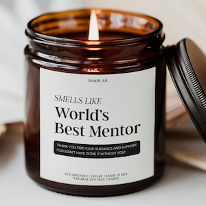 May include: A lit soy wax candle in a brown glass jar with a black lid. The label reads "Smells Like World's Best Mentor" with a thank you message. The candle is eco-friendly, vegan, and made in the USA.
