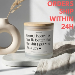 May include: A white candle with a wooden lid, featuring the text "mom, I hope this smells better than the shit I put you through." The candle is on a marble-like surface, with a vase and dried plant in the background.