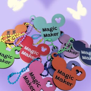 May include: A collection of colorful keychains in the shape of a mouse head, each with the words "Magic Maker" printed on them. The keychains are in various colors, including red, orange, pink, and blue, and are attached to beaded chains.