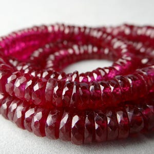 May include: Close-up of a strand of faceted, deep red gemstones. The beads are strung together, forming a spiral shape. The gemstones have a high luster and reflect light, creating a sparkling effect. The background is a soft, neutral color.