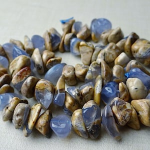 May include: A necklace made of polished, irregular-shaped stones. The stones are a mix of light blue and tan, with variations in color and texture. The necklace is arranged in a circular pattern, suggesting it is a choker or a short necklace.