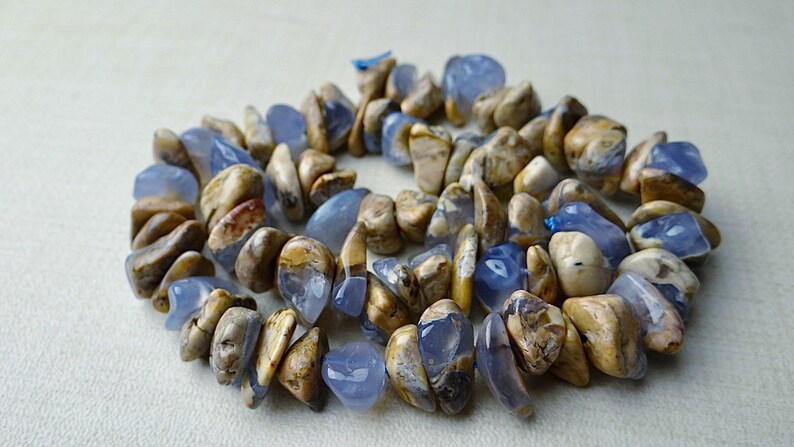 May include: A necklace made of polished, irregular-shaped stones. The stones are a mix of light blue and tan, with variations in color and texture. The necklace is arranged in a circular pattern, suggesting it is a choker or a short necklace.