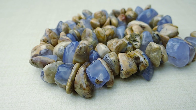 May include: A close-up of a bracelet made of polished, irregular-shaped stones. The stones are a mix of light blue and tan colors. The bracelet is lying on a light-colored surface.