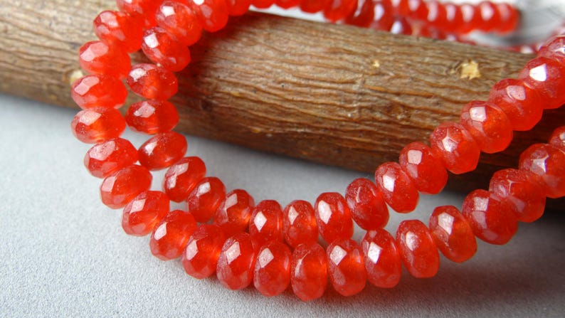 May include: Close-up of a strand of faceted, vibrant red beads. The beads are strung together, creating a necklace or bracelet. The beads have a sparkling, gem-like quality, reflecting light. The background includes a wooden element.