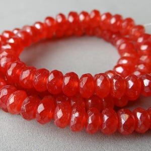 May include: Strands of faceted, vibrant red beads. The beads are strung together, forming a necklace or bracelet. The beads have a polished, glossy appearance, reflecting light. The background is a soft, neutral gray.