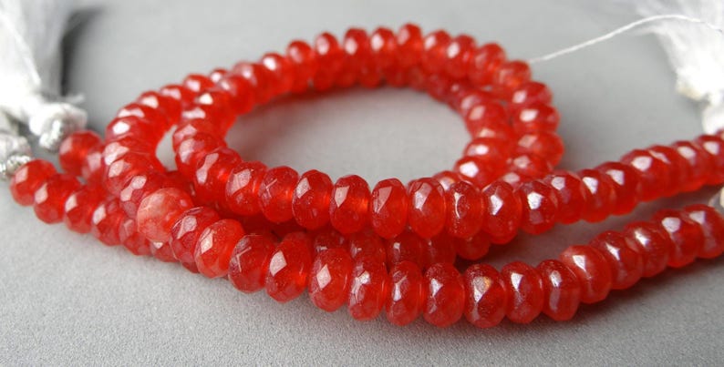 May include: Strands of faceted, vibrant red beads. The beads are strung together, forming a necklace or bracelet. The beads have a polished, glossy appearance, reflecting light. The background is a soft, neutral gray.