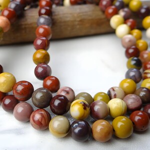 May include: Strands of round gemstone beads in shades of red, yellow, brown, and cream. The beads are strung together, creating a necklace or bracelet. The beads are approximately 0.5 inches in diameter. The background is a light color.